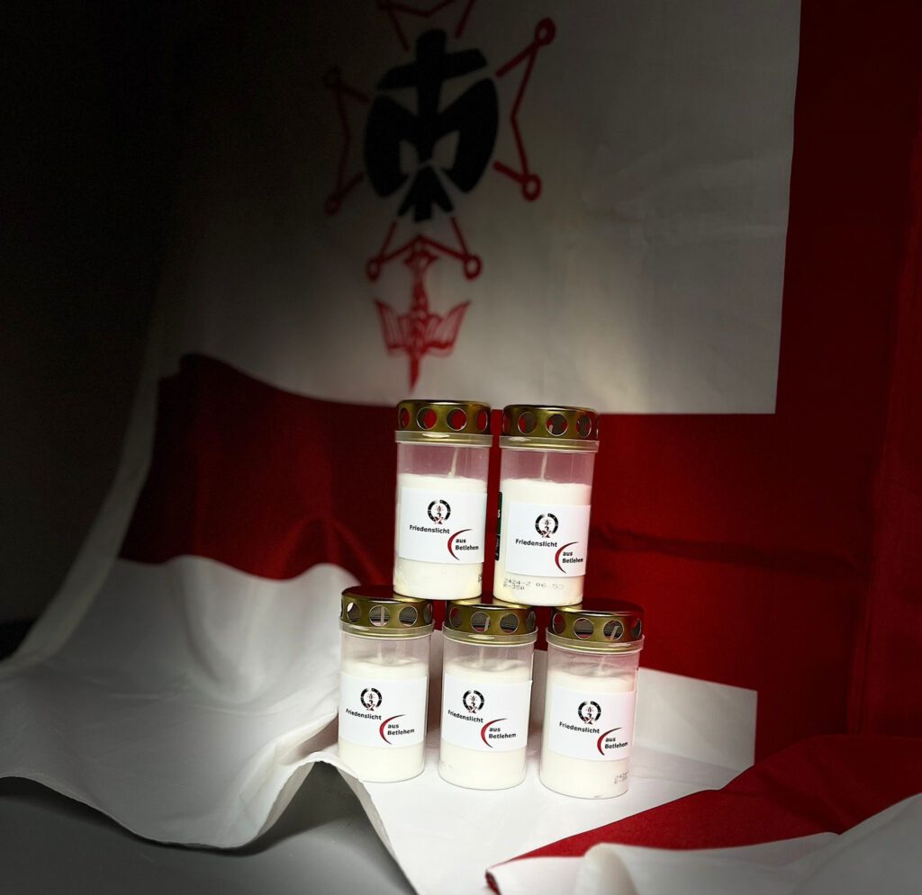 Im Vordergrund werden 5 Kerzen mit dem Friedenslichts Logo die in pyramidenförmig gestäbelt sind gezeigt. Im Hintergrund befindet sich ein Banner von der Pfadfinder der DPSG Warndt-Scouts. 